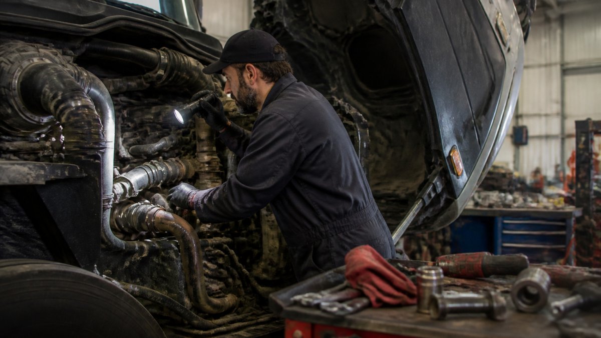 A fleet mechanic in work clothes inspecting a heavy-duty truck engine bay with a flashlight in a service garage, diagnostic tools on a nearby bench